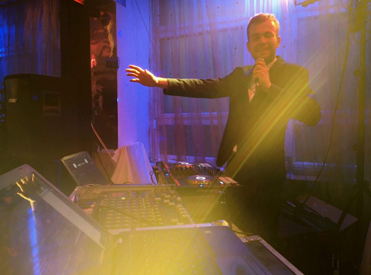 Mężczyzna w garniturze z mikrofonem na tle sprzętu DJ-skiego i kolumny głośnikowej, oświetlony niebieskim i pomarańczowym światłem.