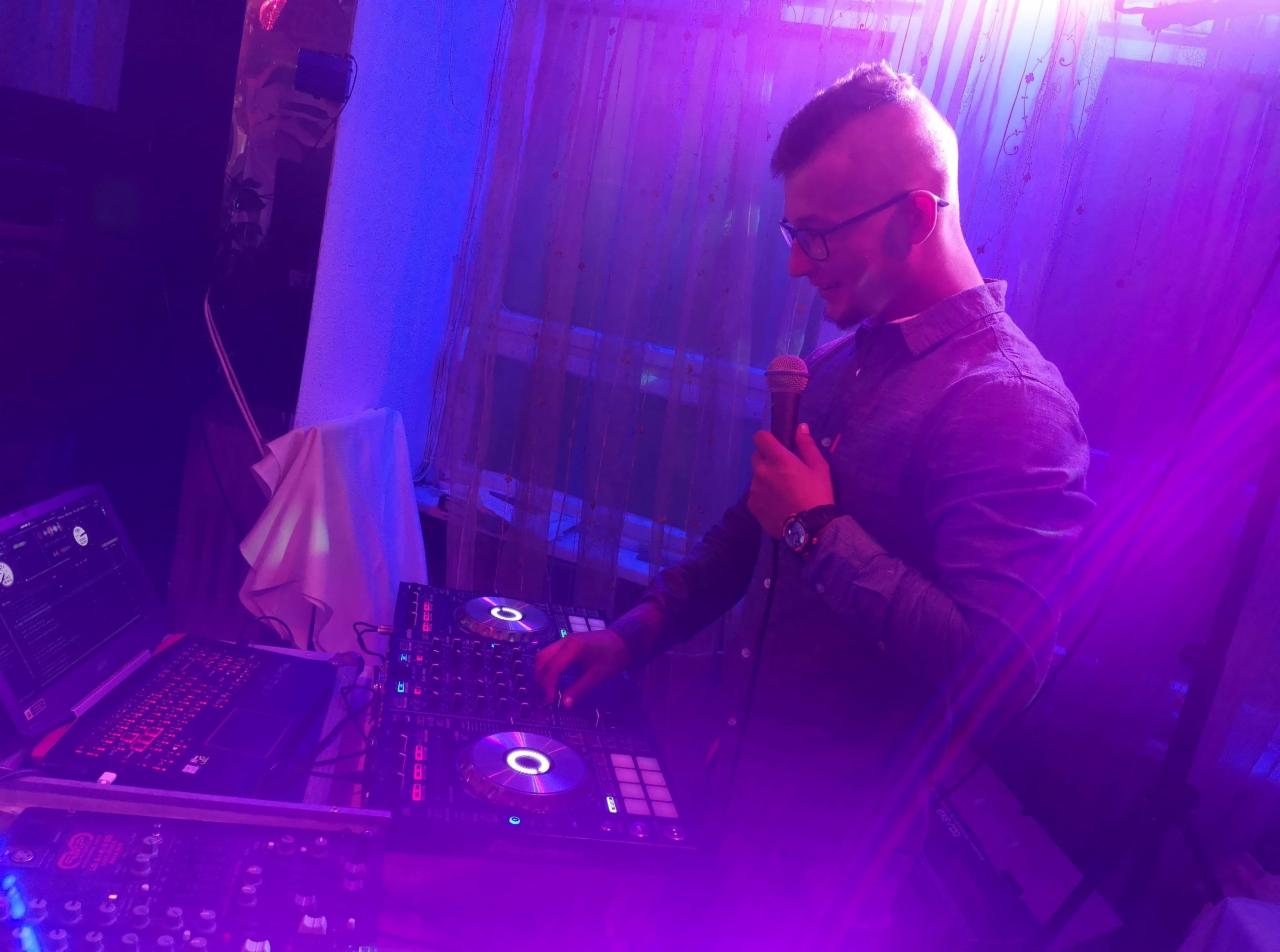 DJ w okularach, z mikrofonem, obsługujący konsoletę DJ-ską oświetloną fioletowym światłem, widoczny laptop z podświetlaną klawiaturą