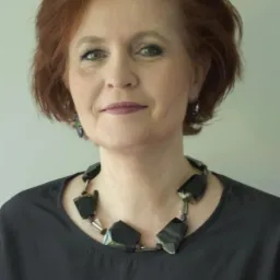 Portret kobiety z rudymi włosami, ubrana w czarną bluzkę i masywny naszyjnik z czarnych kamieni, uśmiechająca się delikatnie.