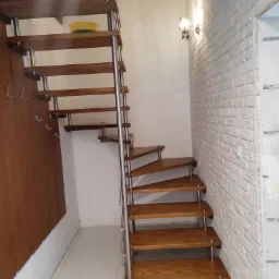Drewniane schody spiralne z metalowymi wspornikami w Białymstoku, prowadzące do góry, obok ściany z białej cegły i drewnianej szafy z wieszakami.