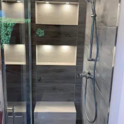 Nowoczesna kabina prysznicowa z szarymi płytkami imitującymi drewno, wbudowanymi półkami z oświetleniem i siedziskiem z tego samego materiału.