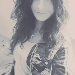 Portret młodej kobiety z kręconymi włosami, w koszuli w kratę i z wisiorkiem z krzyżykiem, na rozmytym tle.