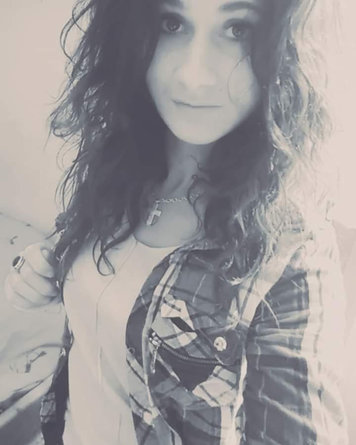 Portret młodej kobiety z kręconymi włosami, w koszuli w kratę i z wisiorkiem z krzyżykiem, na rozmytym tle.