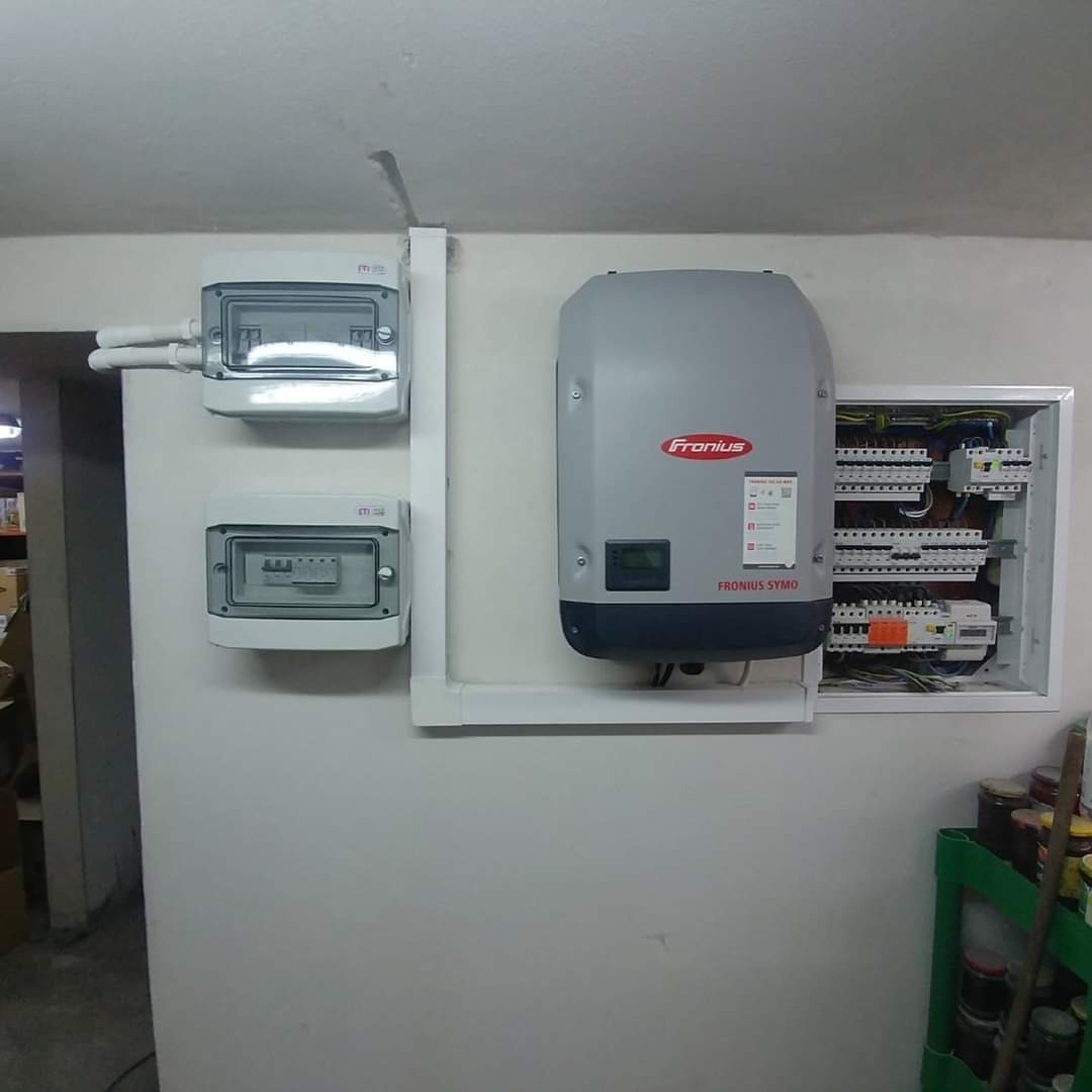 Szara obudowa falownika Fronius Symo, dwie skrzynki elektryczne i otwarta skrzynka rozdzielcza z okablowaniem, zamontowane na ścianie w pomieszczeniu w Łodzi.