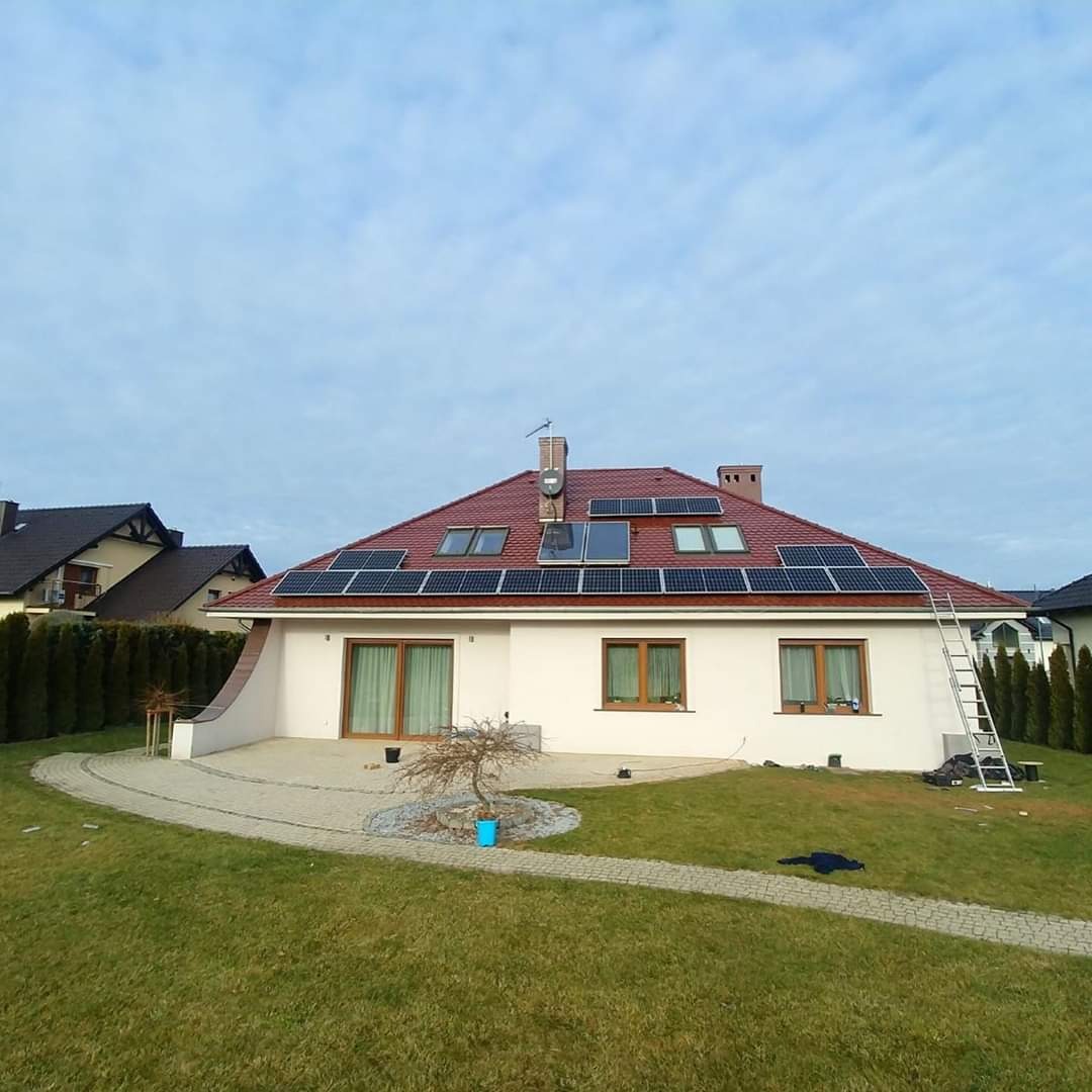 Dom jednorodzinny z czerwoną dachówką i panelami słonecznymi na dachu, drabina oparta o ścianę, trawnik z krzewami, błękitne niebo.