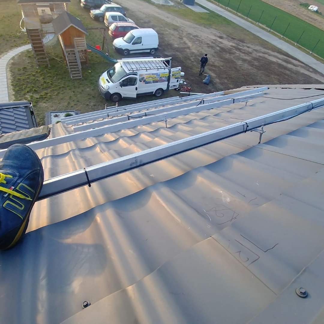 Montaż aluminiowej konstrukcji wsporczej na falistej blasze dachu, widok z góry z butem roboczym w kadrze, w tle samochód dostawczy i plac zabaw.