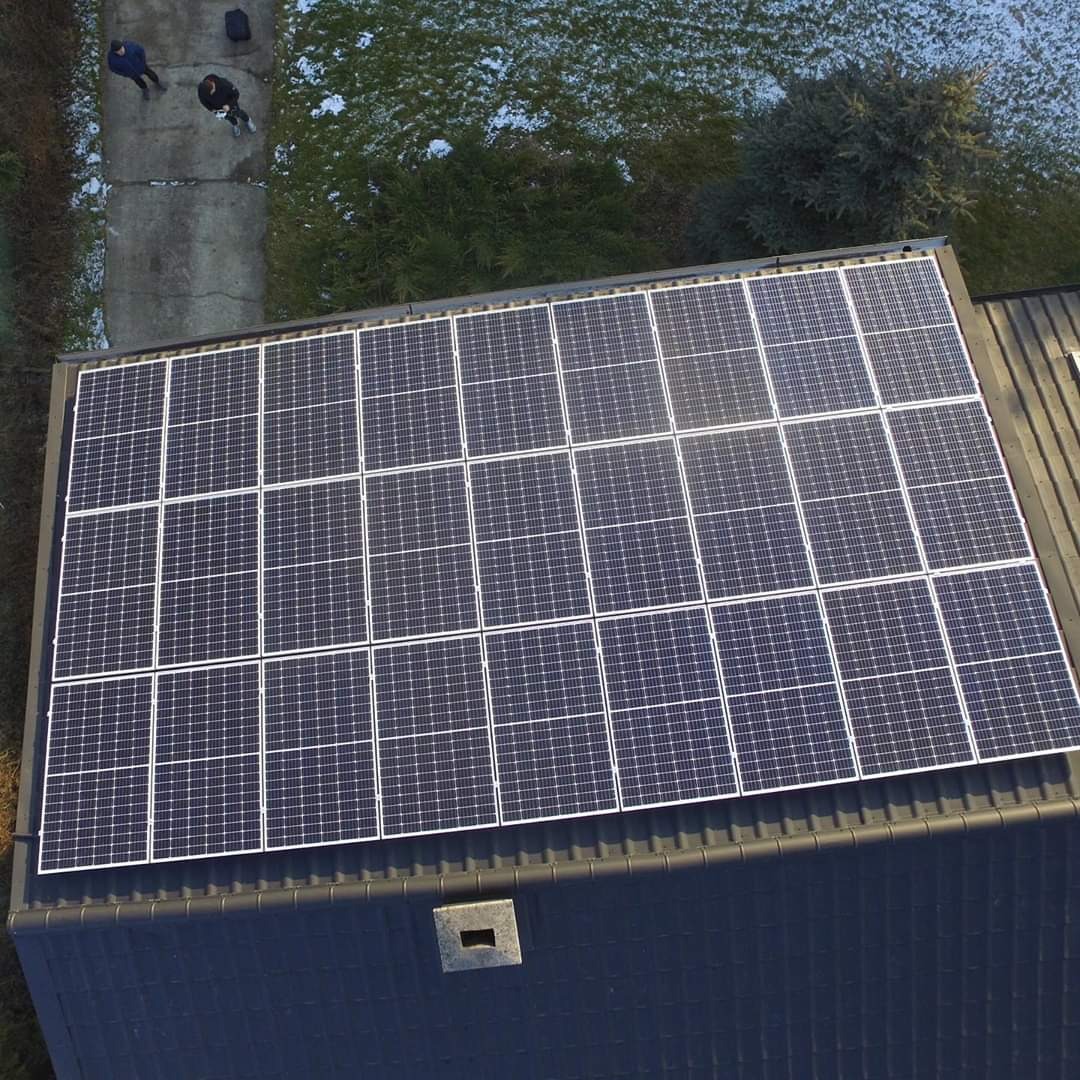 Widok z góry na panel słoneczny na ciemnym dachu domu, z fragmentem chodnika i dwoma osobami w tle, częściowo pokrytym śniegiem.