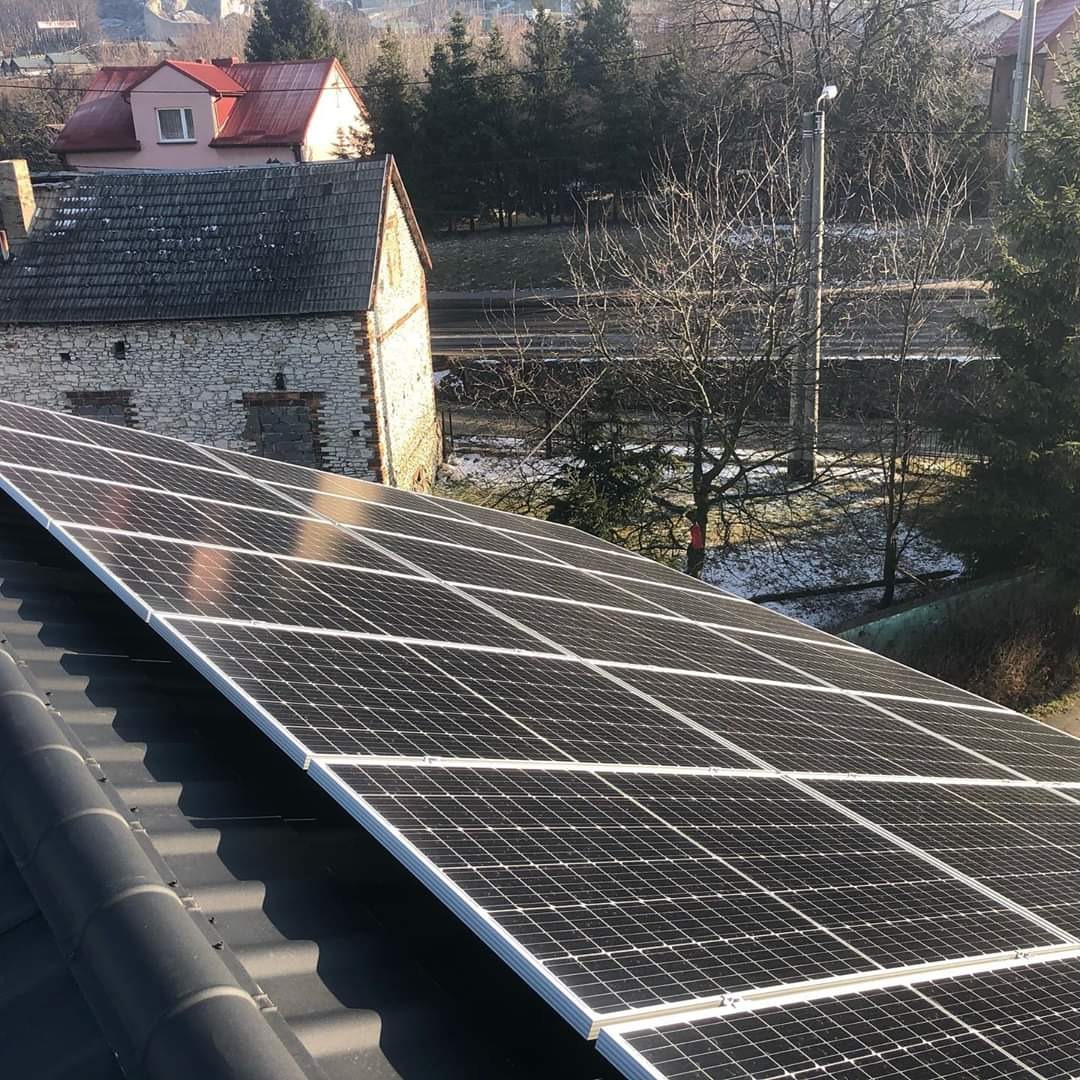 Panel fotowoltaiczny na dachu domu z widokiem na budynek gospodarczy z kamienia i drzewa w tle.