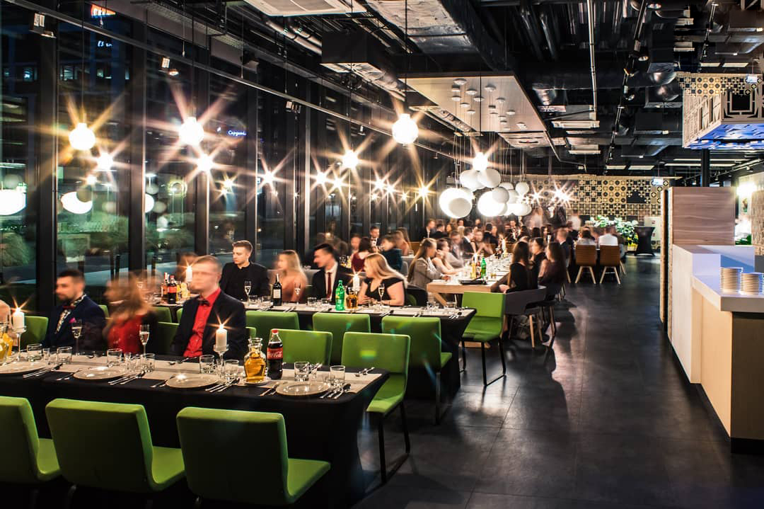 Uroczysta kolacja firmowa w przestronnym wnętrzu z wysokimi oknami, długie stoły nakryte czarnymi obrusami, zielone krzesła, świece i dekoracyjne oświetlenie tworzą elegancką atmosferę.