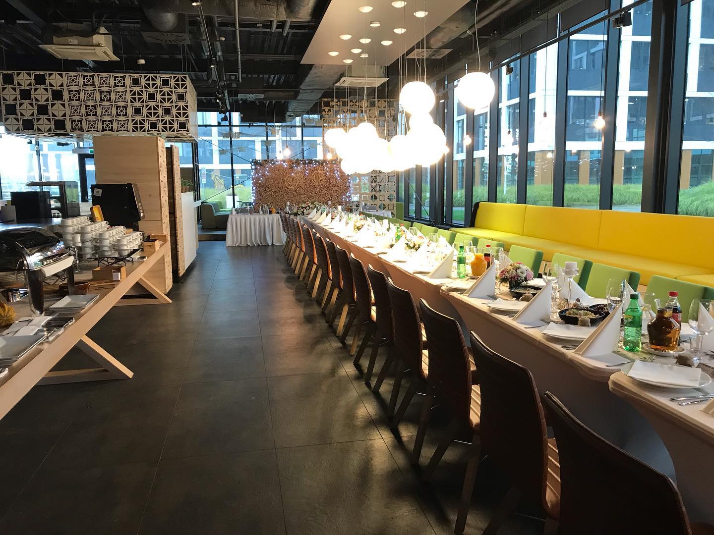 Długi stół z nakryciami i dekoracjami kwiatowymi w nowoczesnej przestrzeni eventowej z dużymi oknami i żółtą sofą w tle, widoczny bufet z potrawami.