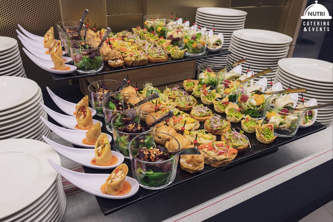 Dwa poziomy zróżnicowanych przekąsek na czarnych tacach: roladki na białych łyżkach, sałatki w szklankach i mini kanapki, obok stosy białych talerzy, w tle logo firmy cateringowej.