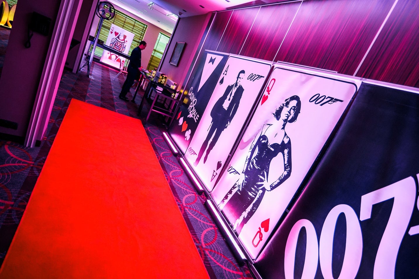 Korytarz z czerwonym dywanem i dekoracjami w stylu Jamesa Bonda: duże karty do gry z sylwetkami postaci, oświetlenie w odcieniach fioletu i różu, w tle osoba w garniturze przy stole.