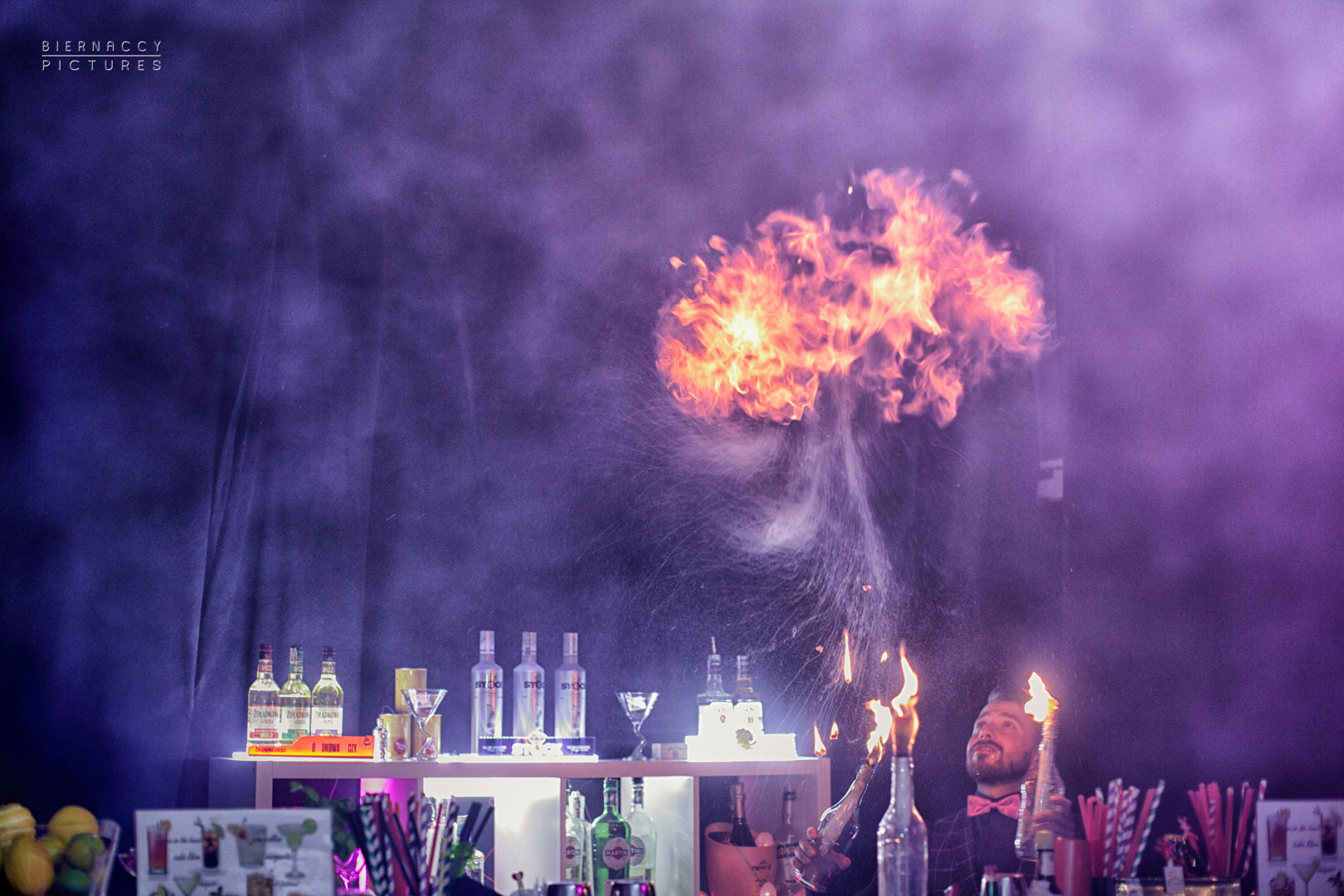 Barman w muszce żongluje płonącymi butelkami, tworząc efektowny pokaz ognia nad barem z drinkami.
