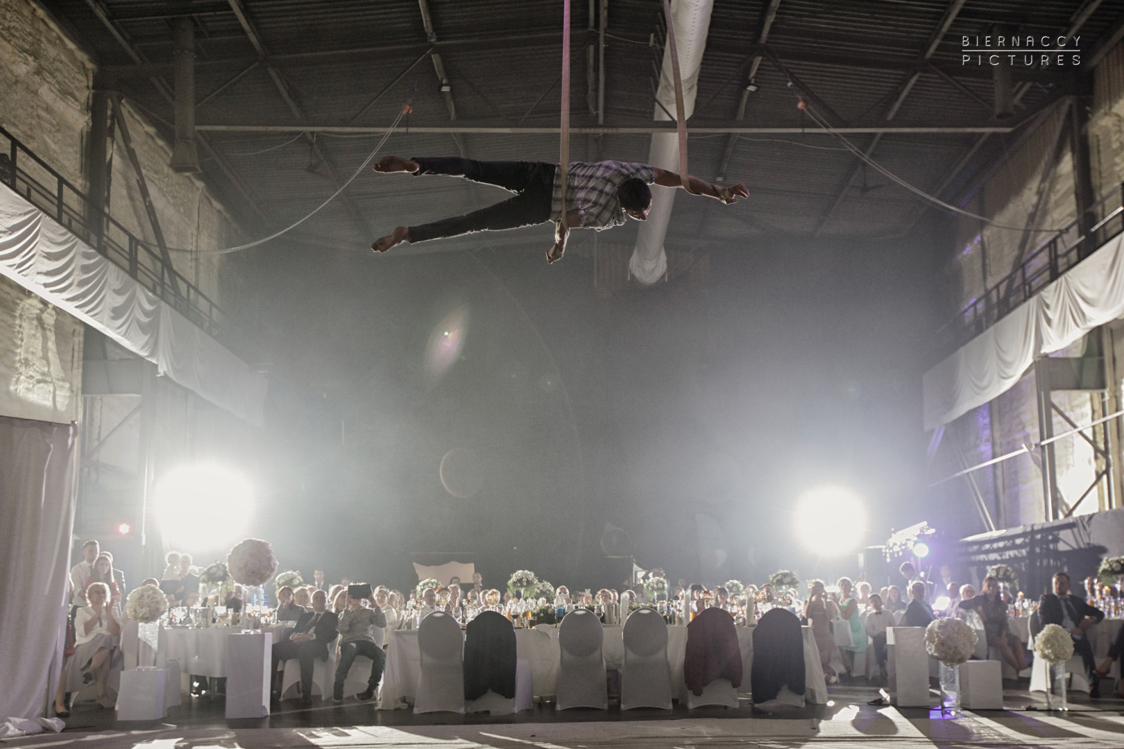 Akrobata powietrzny w koszuli w kratę wykonuje pokaz nad stołami z gośćmi na imprezie w hali z surową konstrukcją, oświetlony reflektorami.