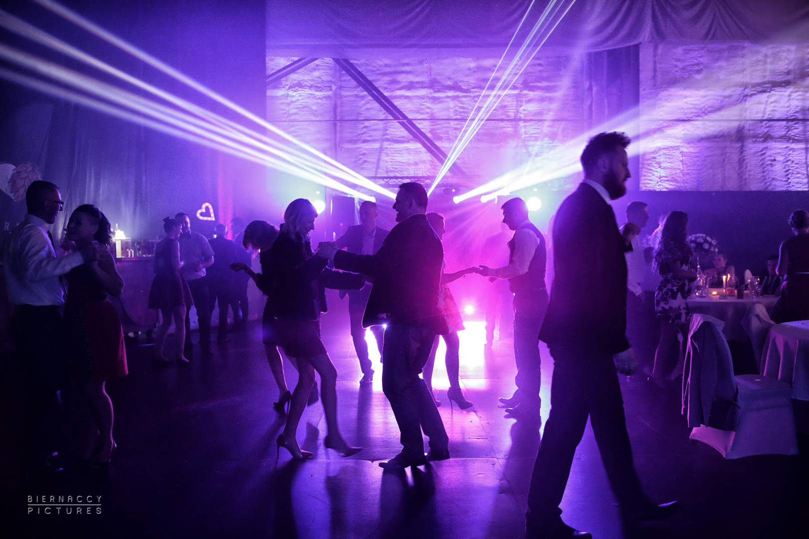 Uczestnicy w ruchu na parkiecie podczas imprezy, oświetleni fioletowymi laserami i reflektorami, widoczny stół z dekoracjami w tle.