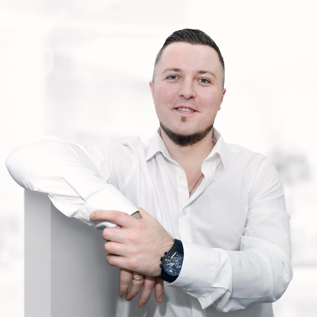 Portret mężczyzny z brodą i w białej koszuli, opierającego się o biały element, z zegarkiem na ręce, na rozmytym, jasnym tle, Wrocław.