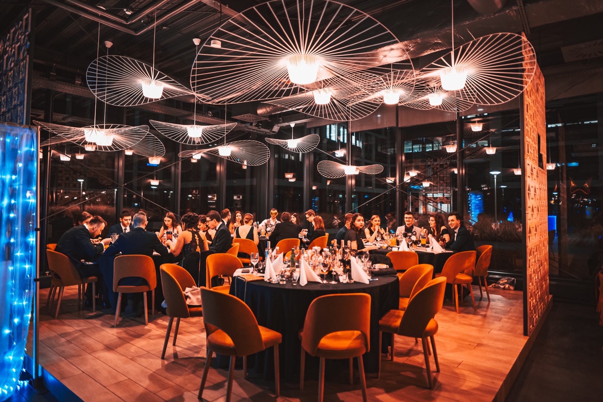 Uroczysta kolacja firmowa w nowoczesnej sali z dekoracyjnymi lampami, okrągłe stoły z czarnymi obrusami, pomarańczowe krzesła, uczestnicy w strojach wieczorowych.