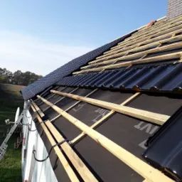 Montaż czarnej dachówki ceramicznej na łatach pokrytych czarną membraną, widoczny komin i drabina oparta o ścianę budynku.