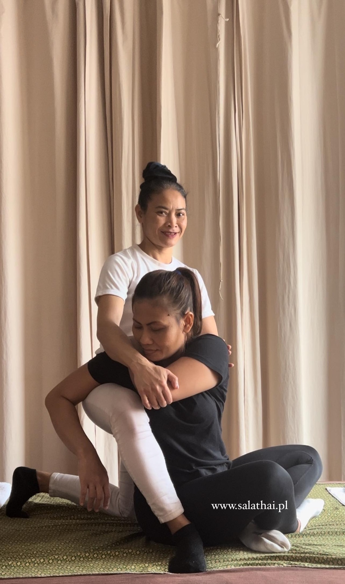Terapeutka w białej koszulce i spodniach, używa kolana do delikatnego ucisku na plecach klientki w czarnej koszulce, podczas sesji na zielonej macie przed beżową zasłoną.