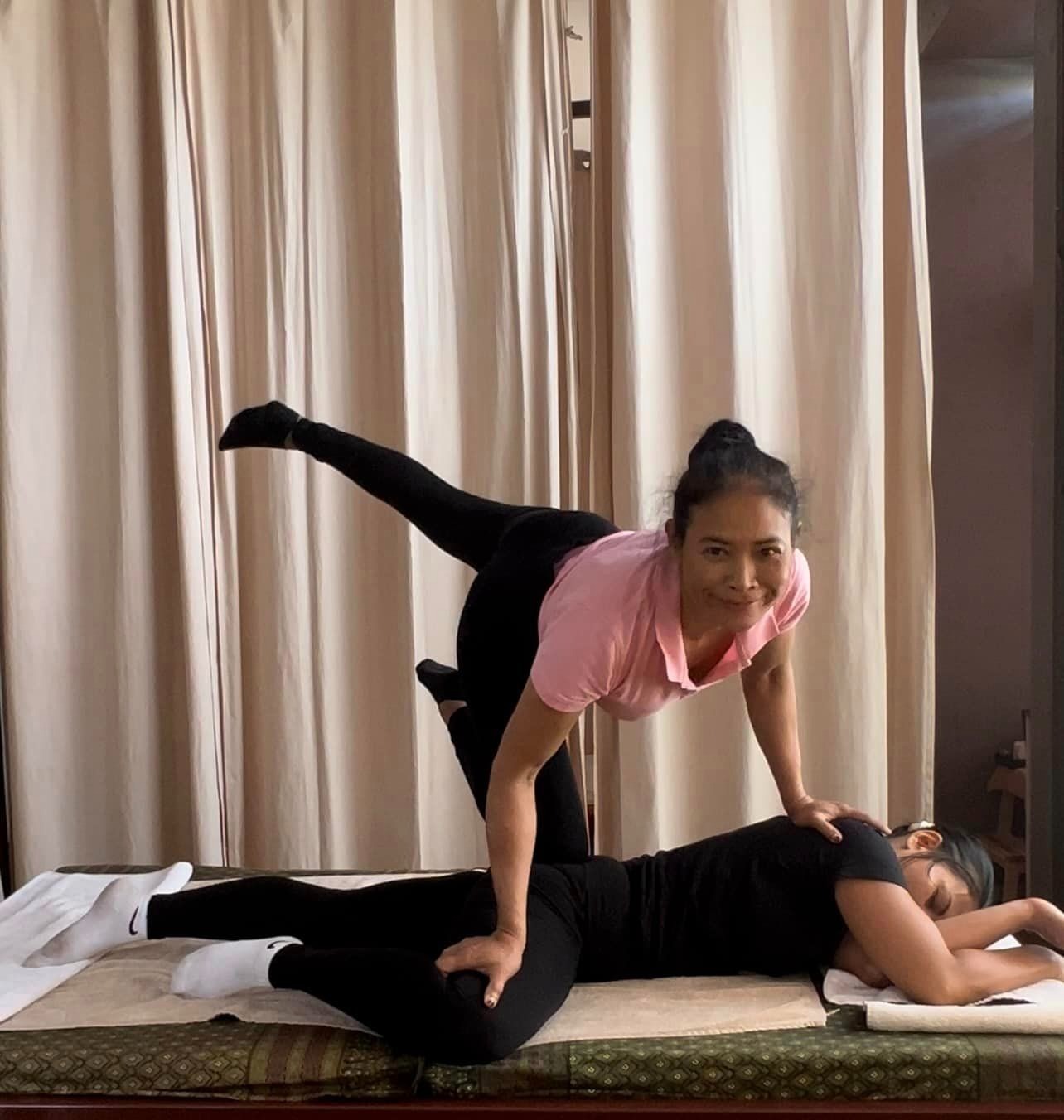 Kobieta w różowej koszulce wykonuje tradycyjny masaż tajski, opierając się na plecach leżącej osoby i używając nóg do rozciągania, na tle beżowych zasłon.