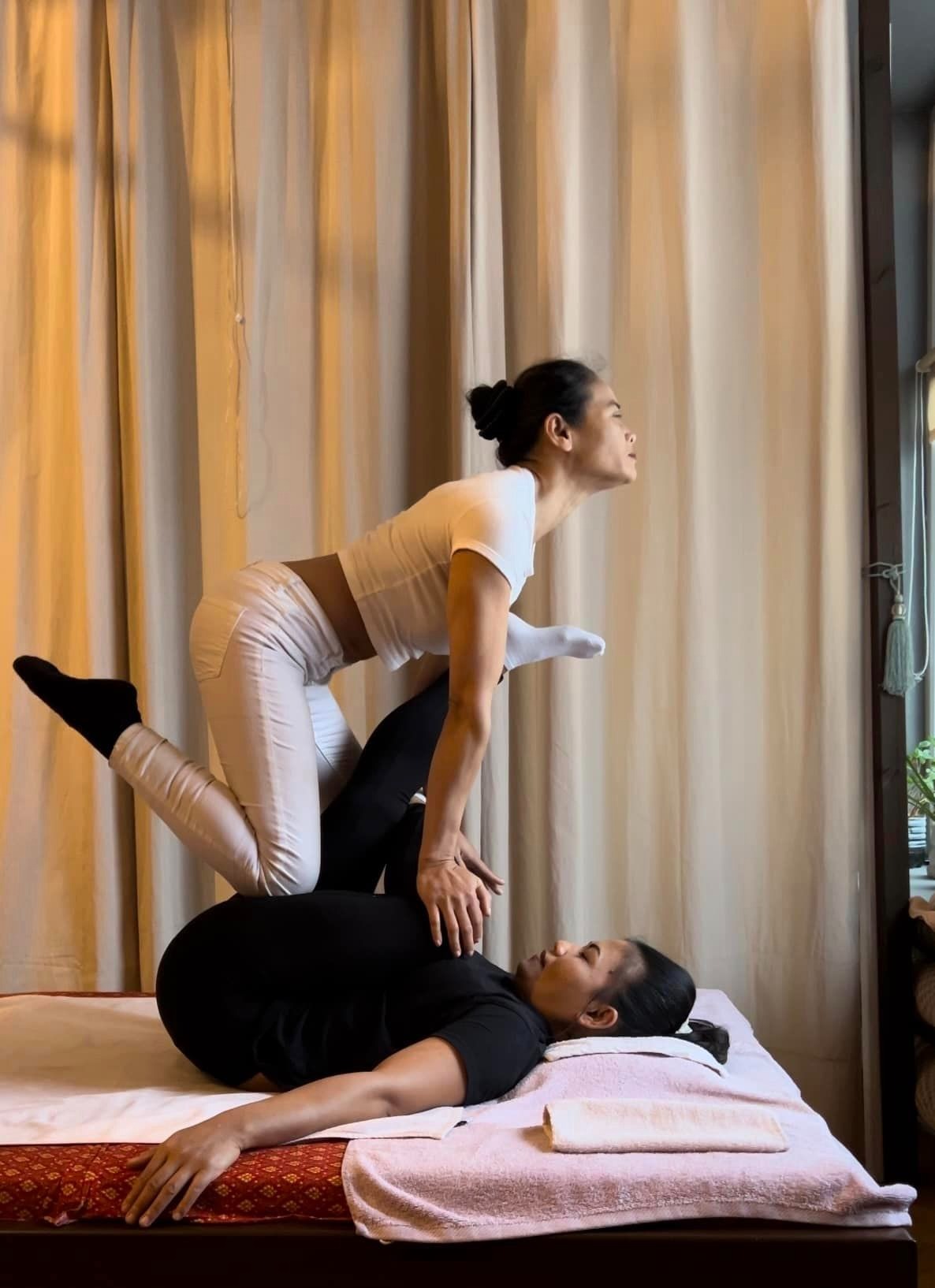 Kobieta w białym stroju wykonuje tajski masaż na leżącej kobiecie w czarnym stroju, wykorzystując stopę do nacisku na plecy, w tle zasłony i okno.