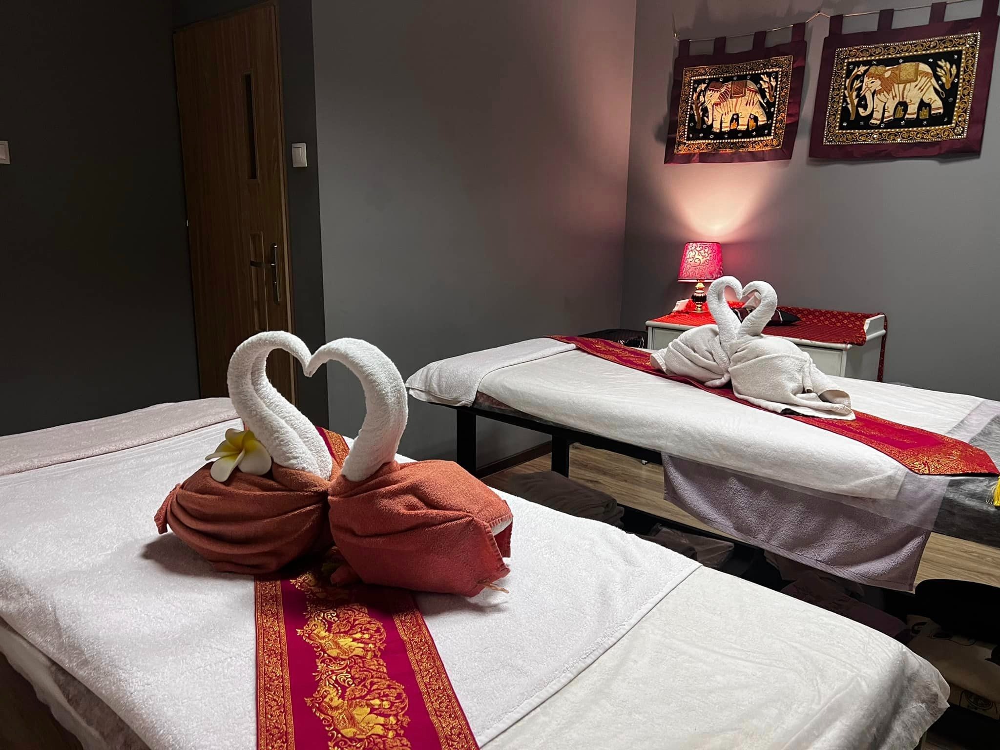 Dwa łóżka do zabiegów w gabinecie, udekorowane ręcznikami uformowanymi w kształt łabędzi z kwiatem, z bordowymi bieżnikami z motywami orientalnymi i obrazami słoni na ścianie.