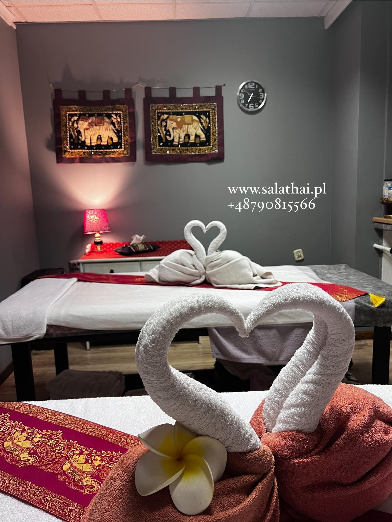 Ustawione łóżka do zabiegów w salonie z tajskim motywem, ręczniki uformowane w kształt łabędzi i serc, dekoracyjne obrazy słoni na ścianie, zegar.