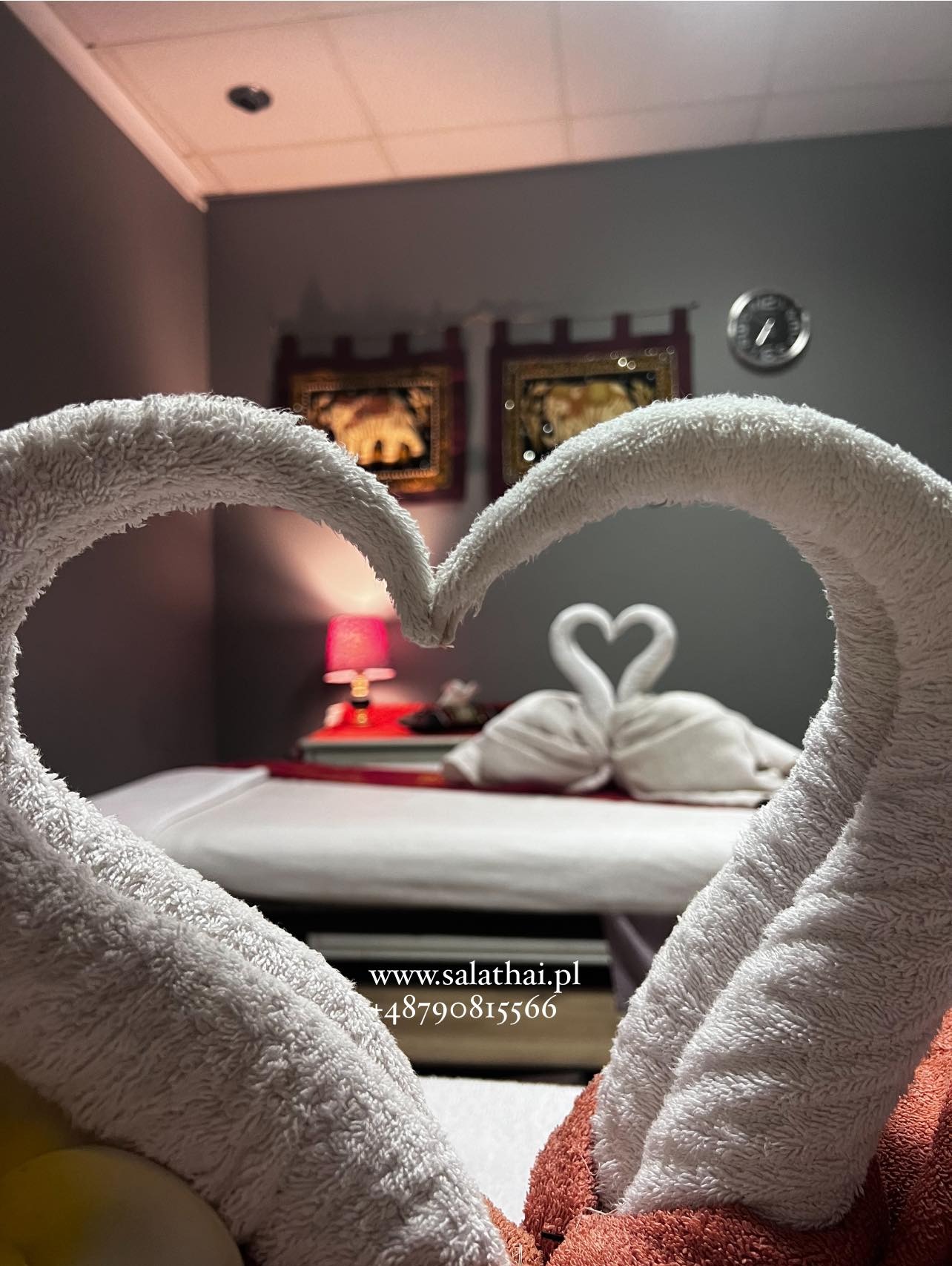 Ułożone w serce białe ręczniki na stole do zabiegów, w tle dekoracja z ręczników uformowanych w łabędzie, przytulne oświetlenie i tajskie obrazy na ścianie.