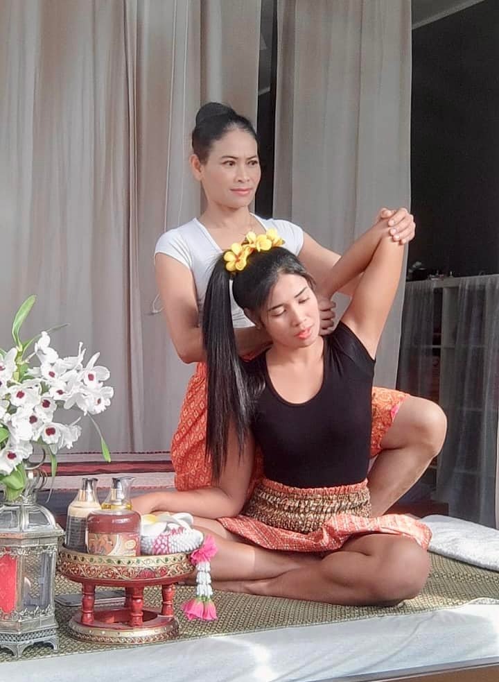Kobieta w trakcie tajskiego rozciągania ramion przez masażystkę w tradycyjnym otoczeniu, z dekoracyjnym stolikiem i kwiatami.