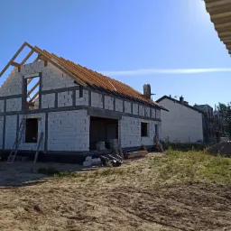 Surowy stan budynku z bloczków betonowych z drewnianą konstrukcją dachu w trakcie budowy, widoczna drabina oparta o ścianę, na drugim planie inny budynek w budowie, słoneczny dzień.