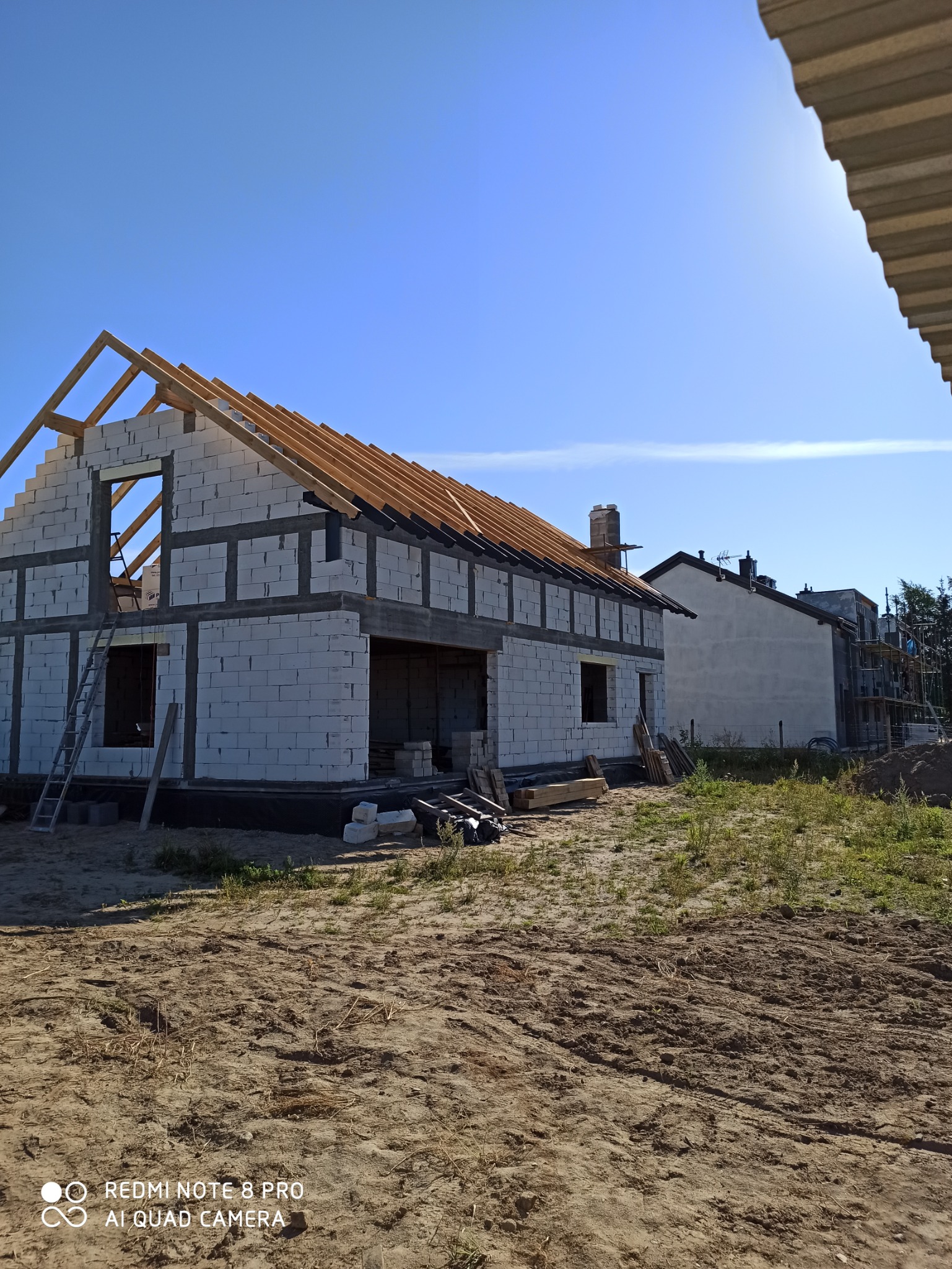 Surowy stan budynku z bloczków betonowych z drewnianą konstrukcją dachu w trakcie budowy, widoczna drabina oparta o ścianę, na drugim planie inny budynek w budowie, słoneczny dzień.