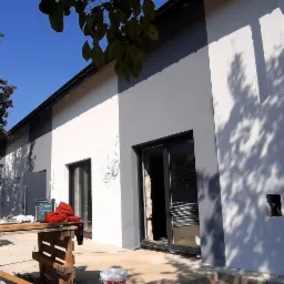 Dwukolorowa elewacja domu w trakcie wykańczania, z widocznymi śladami po ołówku na świeżej, jasnej farbie, oraz narzędzia budowlane i rusztowania w otoczeniu.