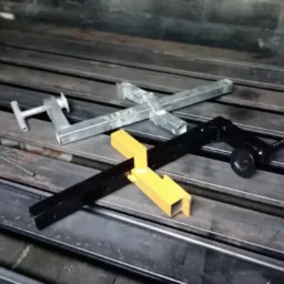 Metalowe elementy konstrukcyjne, w tym spawane profile, w kolorach srebrnym, żółtym i czarnym, leżące na metalowym stole warsztatowym z widocznymi śladami użytkowania.