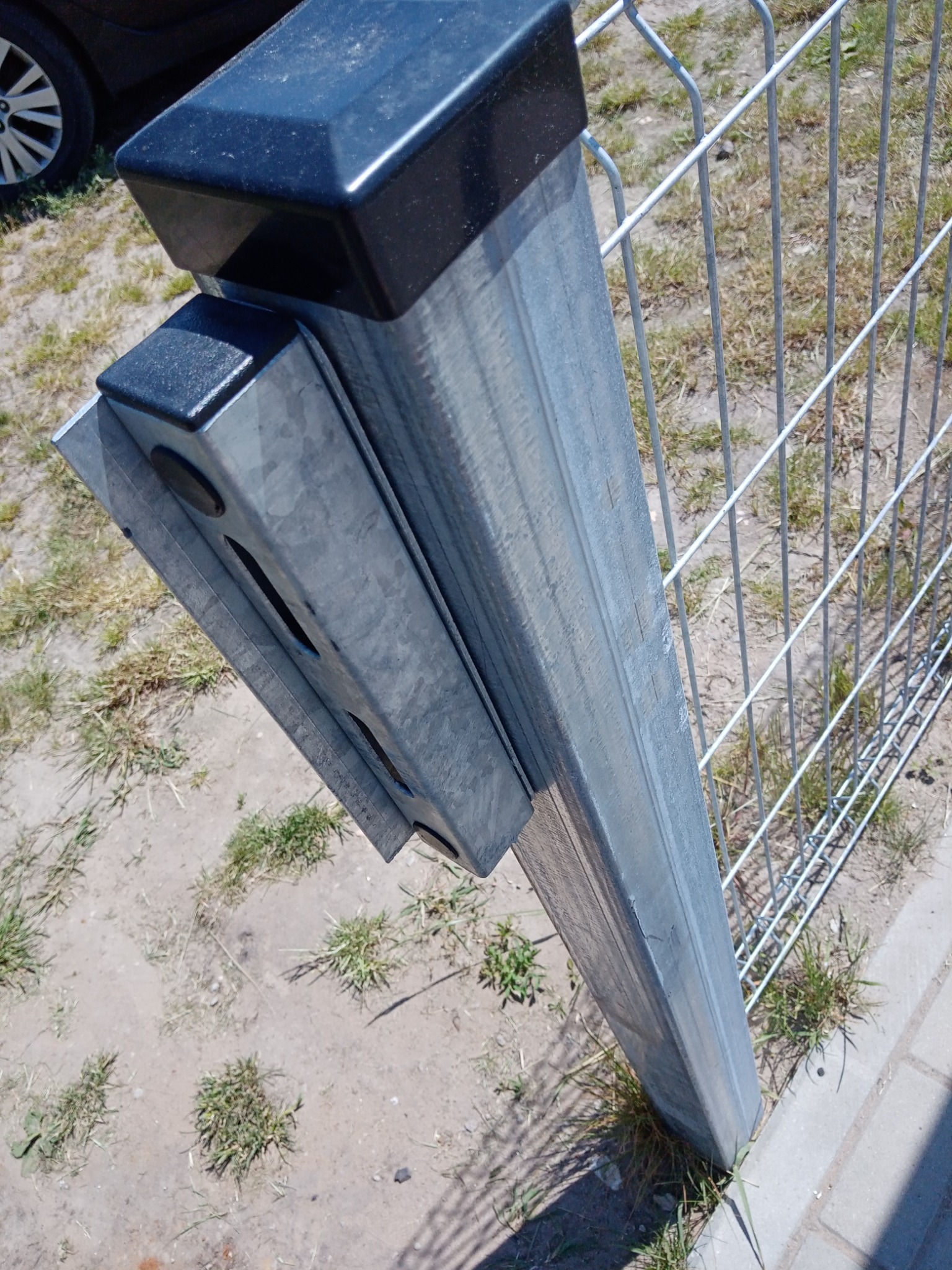 Metalowy słupek ogrodzeniowy z czarnym kapturkiem, przymocowany do siatki, widoczny z bliska na tle trawnika i kostki brukowej.