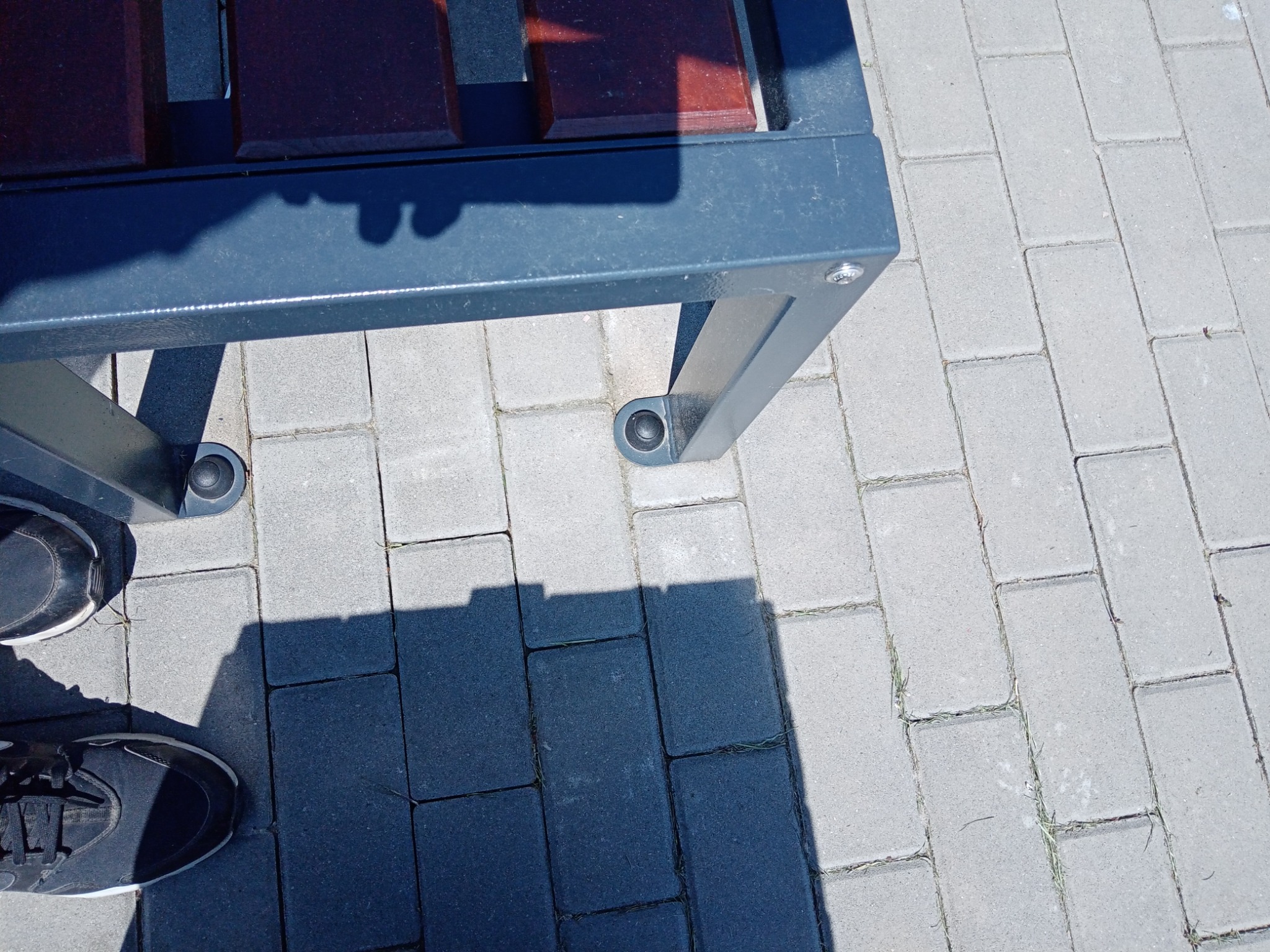 Fragment ławki parkowej z metalową konstrukcją i drewnianym siedziskiem, stojącej na szarej kostce brukowej, z widocznymi butami osoby fotografującej w lewym dolnym rogu.