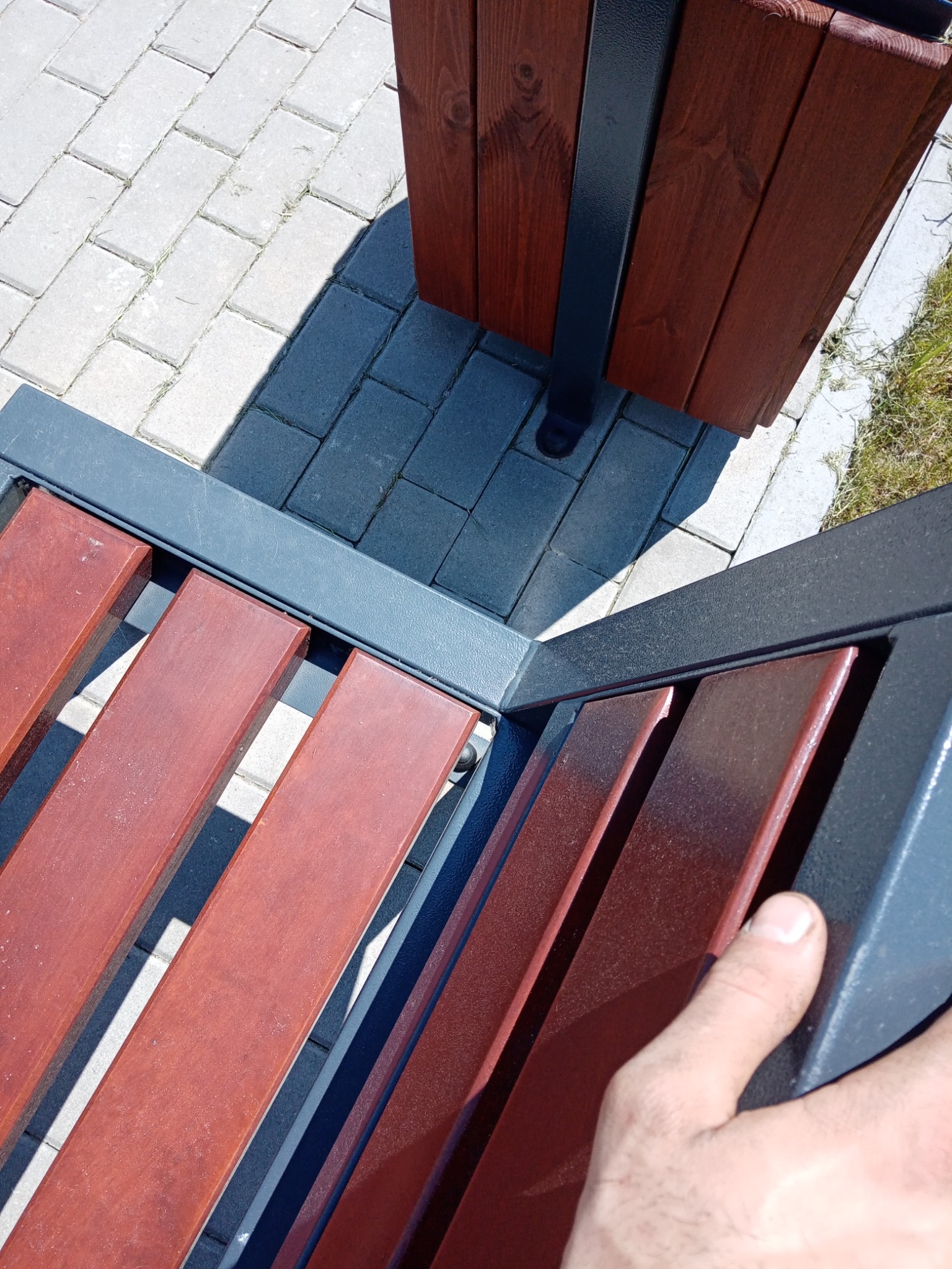 Fragment ławki parkowej z drewnianymi listwami i metalową konstrukcją, obok kosz na śmieci z drewnianą obudową, widoczne fragmenty kostki brukowej i trawy, ujęcie z góry z dłonią w kadrze.