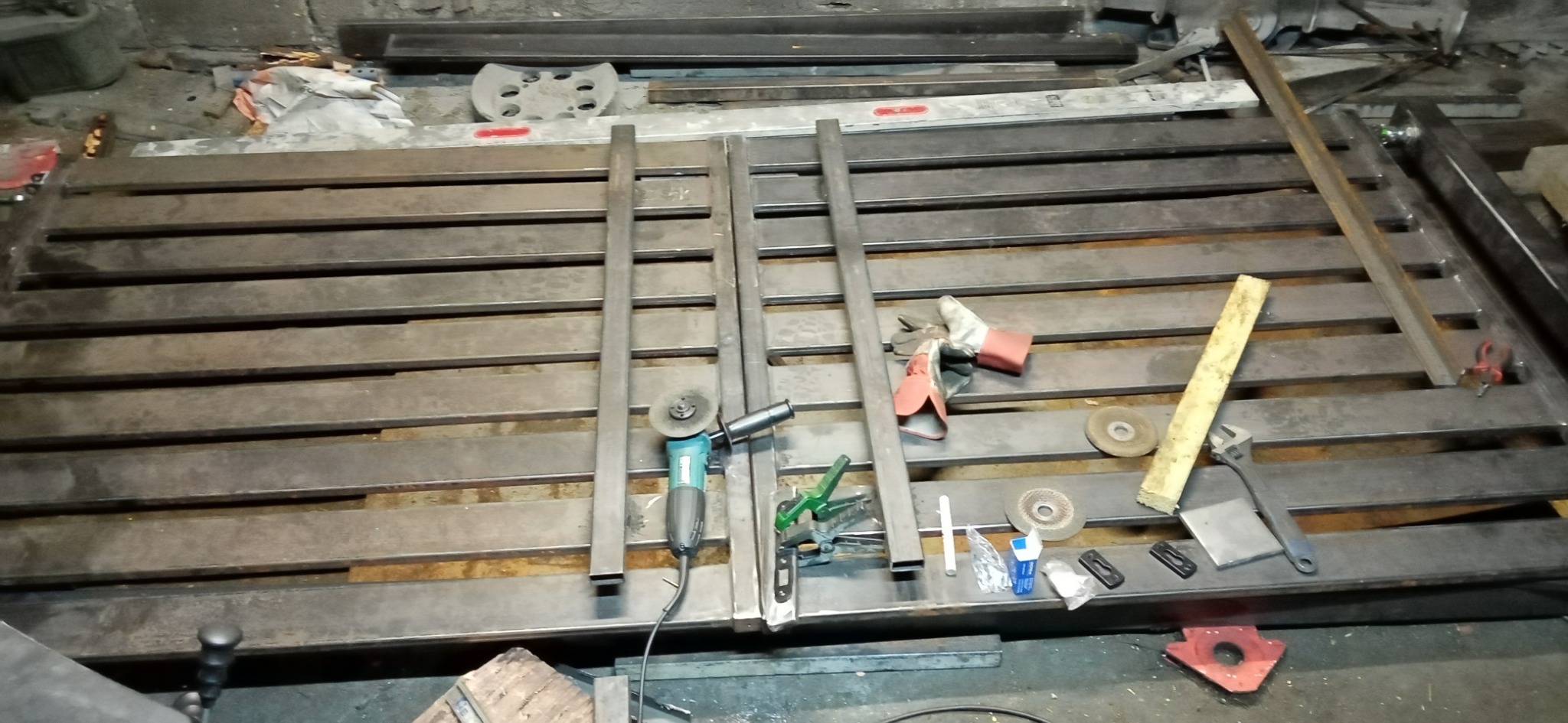 Metalowa konstrukcja bramy w trakcie budowy w warsztacie, widoczne profile, szlifierka kątowa, ściski, rękawice robocze i inne narzędzia.