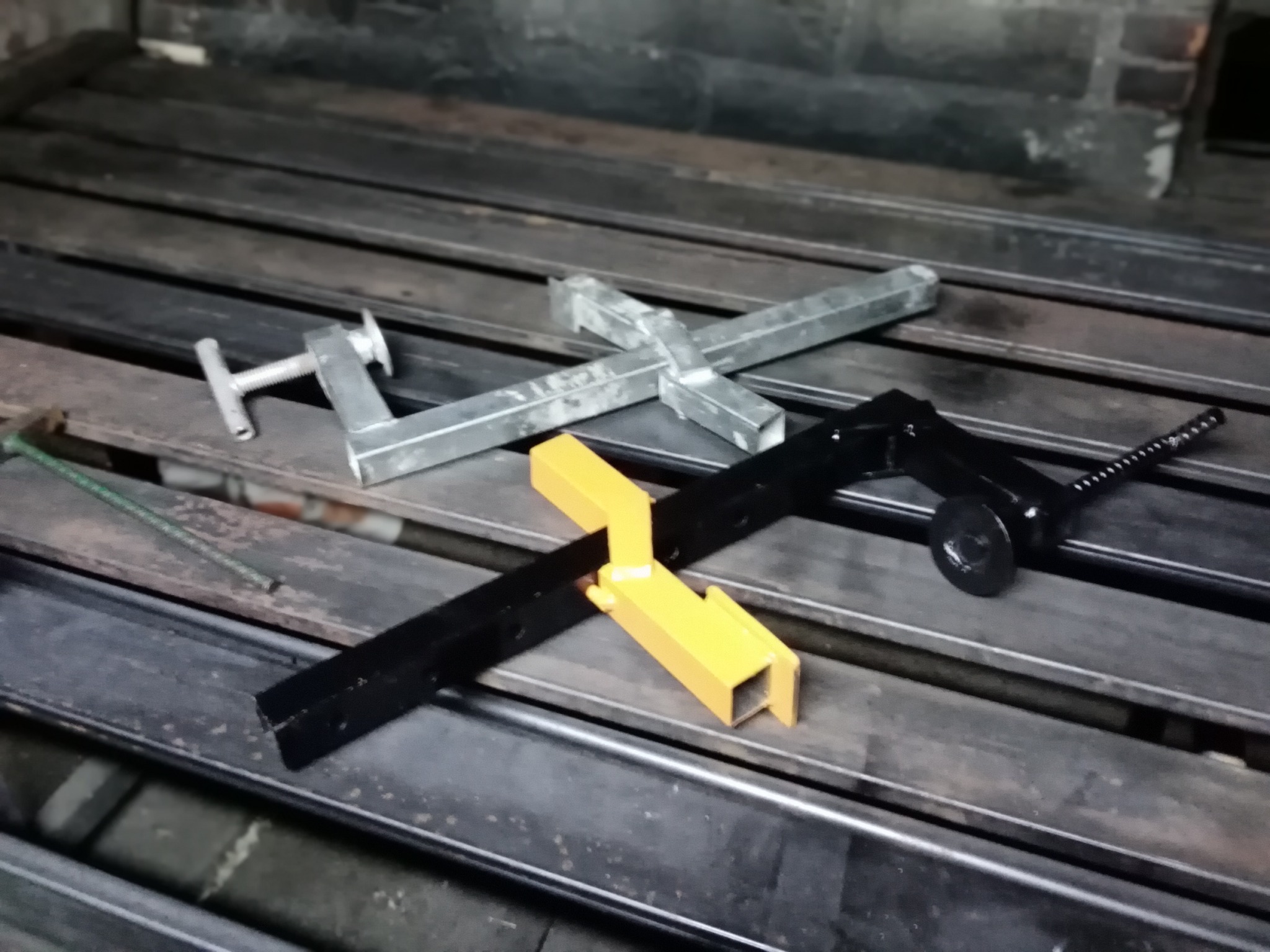 Metalowe elementy konstrukcyjne, w tym spawane profile, w kolorach srebrnym, żółtym i czarnym, leżące na metalowym stole warsztatowym z widocznymi śladami użytkowania.