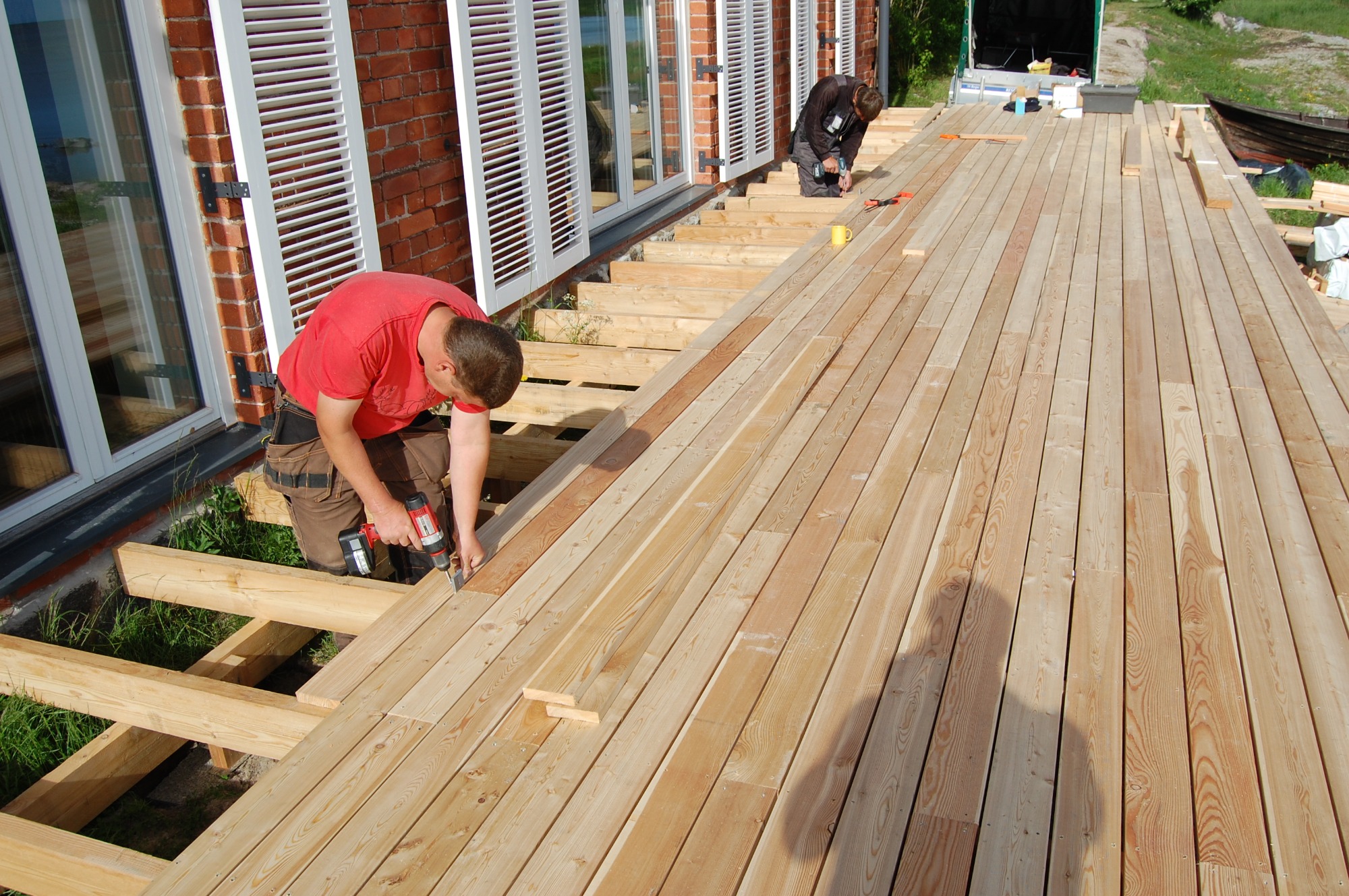 Dwóch mężczyzn montuje drewniany taras przy domu w Tarnogrodzie, używając wkrętarek. Widoczne jasne deski tarasowe, konstrukcja nośna i fragment elewacji budynku z białymi okiennicami.