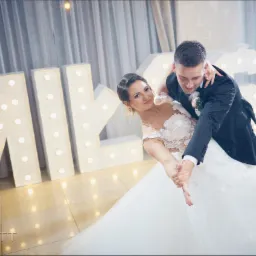 Para młoda tańczy taniec weselny na parkiecie, w tle dekoracyjne, podświetlane litery z napisem.