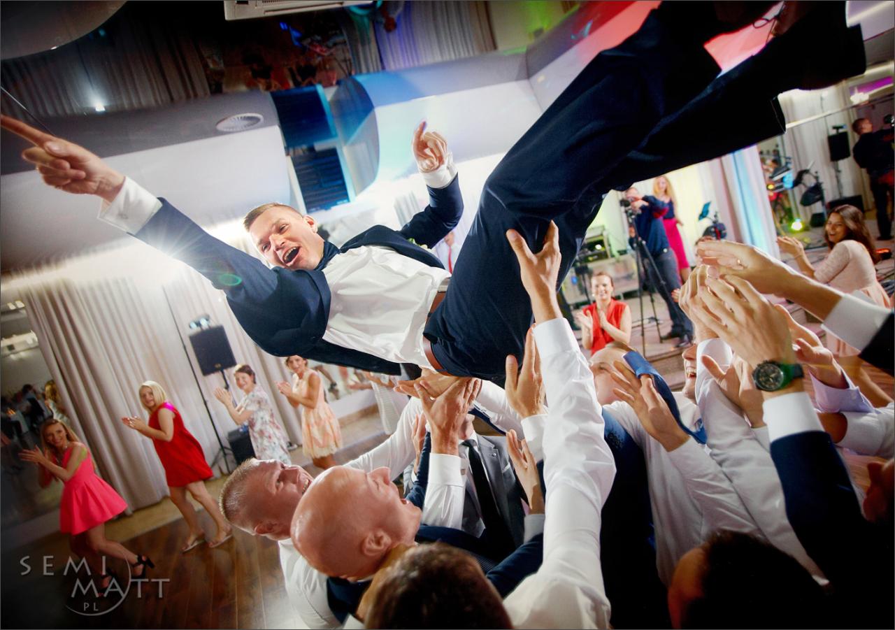 Uśmiechnięty pan młody w garniturze, podrzucany przez gości weselnych podczas zabawy na sali, z widocznym parkietem i oświetleniem imprezowym w tle.
