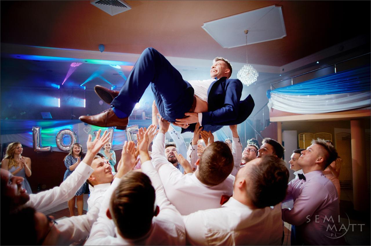 Uśmiechnięty pan młody w niebieskim garniturze podrzucany do góry przez gości weselnych na sali z balonami w kształcie napisu LOVE i dekoracjami w odcieniach błękitu.