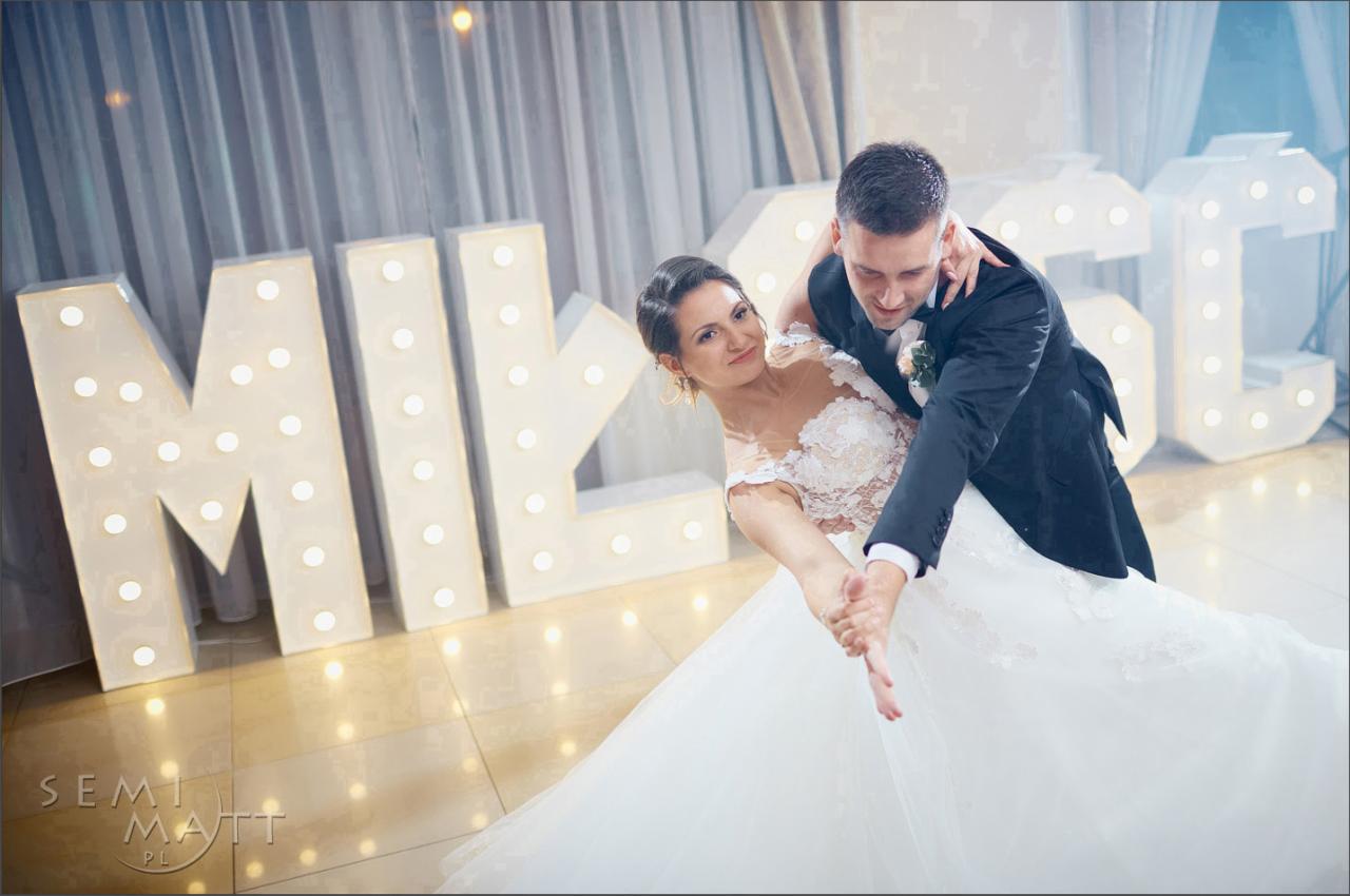 Para młoda tańczy taniec weselny na parkiecie, w tle dekoracyjne, podświetlane litery z napisem.