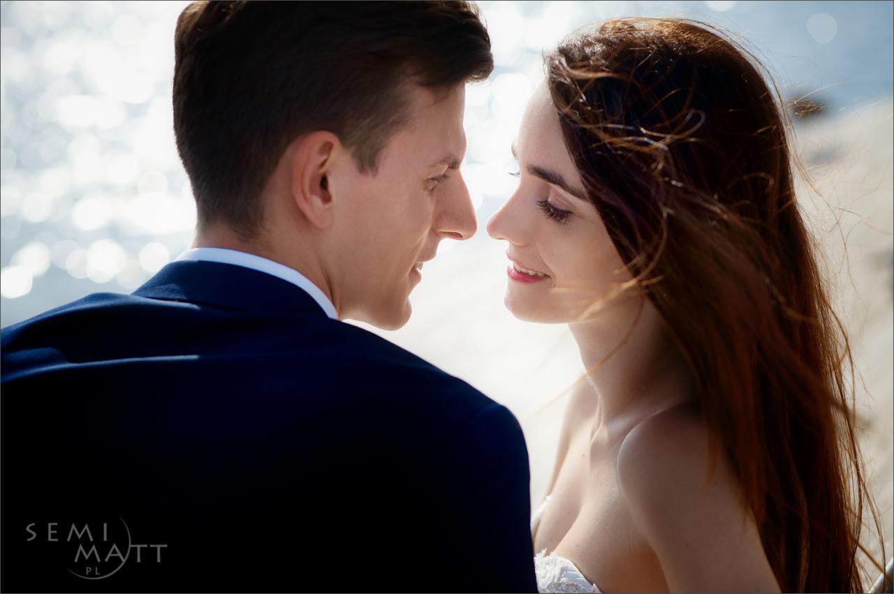 Para młoda patrzy na siebie w profilu, w tle rozmyte światło wody, on w granatowym garniturze, ona z długimi brązowymi włosami, delikatny uśmiech.