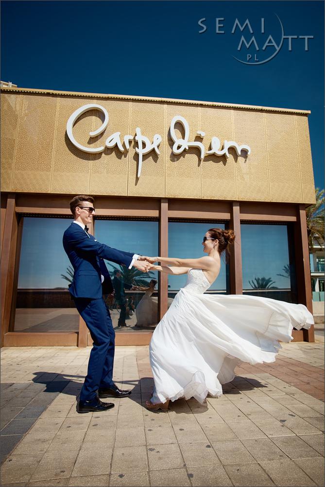 Para młoda w tańcu przed budynkiem z napisem 'Carpe Diem', on w granatowym garniturze i okularach, ona w białej sukni, w tle odbicie w szybach.