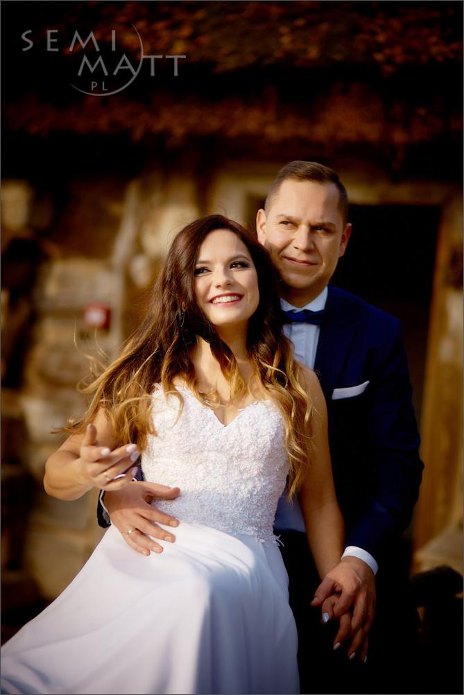 Uśmiechnięta panna młoda w białej sukni z koronkową górą, trzymana w pasie przez pana młodego w granatowym garniturze z muchą, na tle kamiennej ściany.