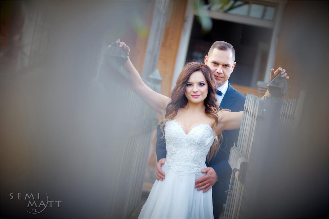 Para młoda w białej sukni i granatowym garniturze pozuje przy drewnianej bramie, z delikatnym efektem rozmycia w pierwszym planie.