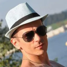 Portret młodego mężczyzny w słomkowym kapeluszu i okularach przeciwsłonecznych na tle rozmytego krajobrazu z jeziorem i zielenią. Ujęcie z bliska, naturalne światło.