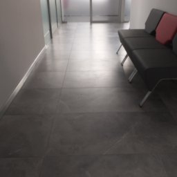 Olkam Dariusz Bylina - Wyłożona szarymi płytkami podłoga w korytarzu biurowym z sofą i przeszklonymi drzwiami w tle.