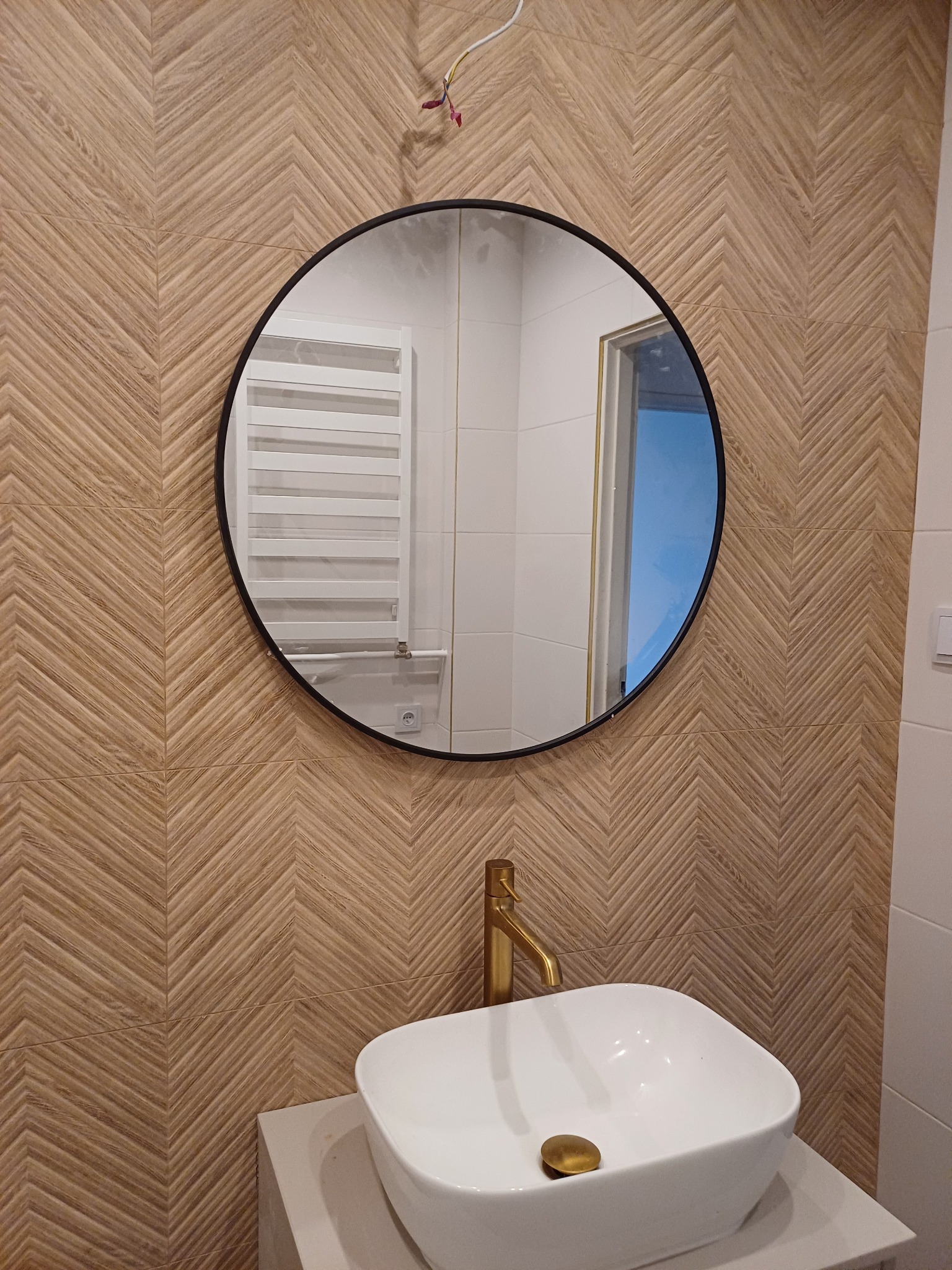 Umywalka nablatowa z baterią w kolorze złotym na tle ściany wyłożonej płytkami imitującymi drewno ułożonymi w jodełkę, okrągłe lustro z czarną ramą odbija grzejnik drabinkowy i fragment otwartych...